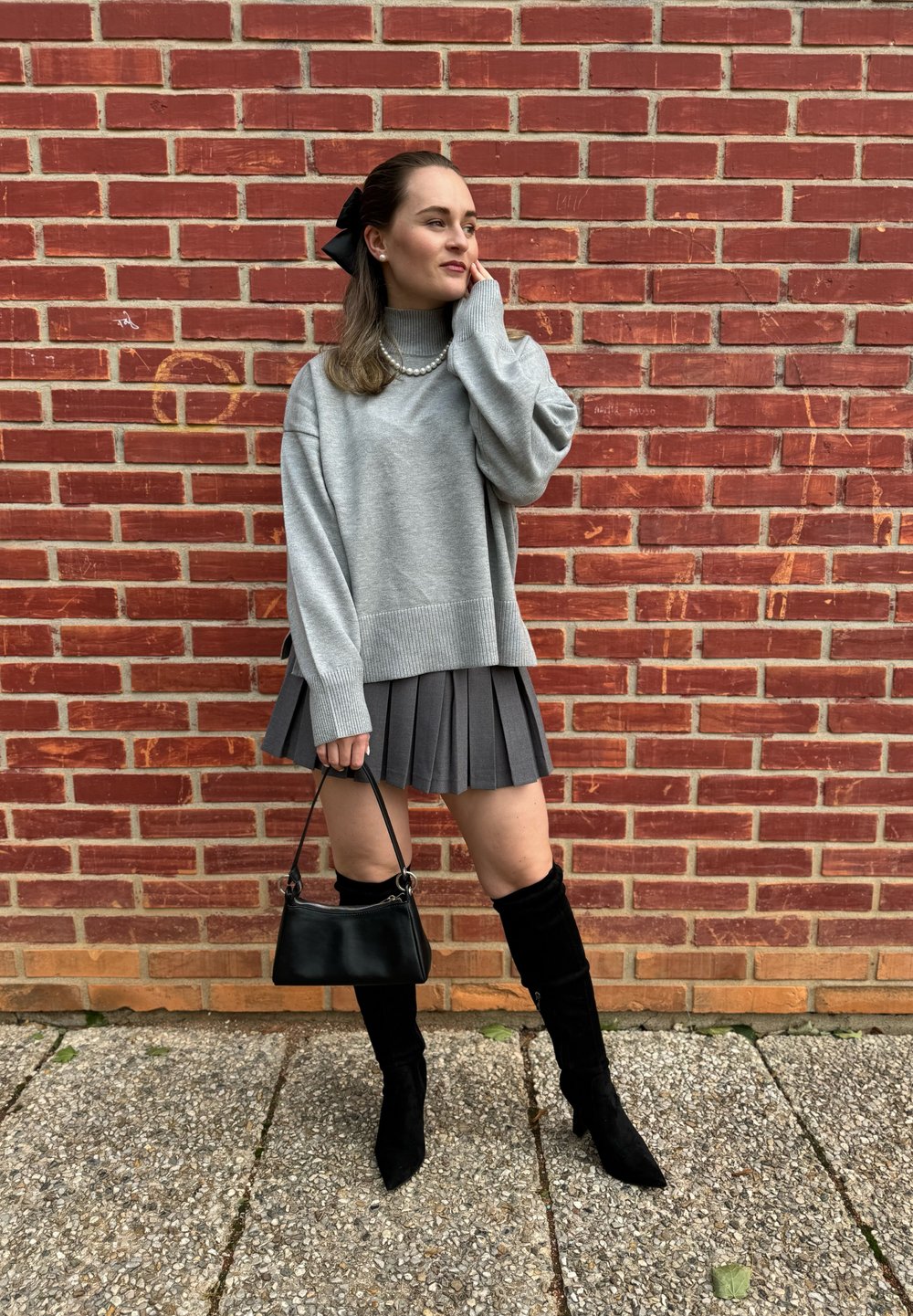 Γκρι oversized πουλόβερ, πλισέ γκρι φούστα, μαύρες ψηλές μπότες και μαύρη τσάντα κατά μήκος ενός τοίχου από τούβλα. Υφές και προσεγμένος σχεδιασμός.