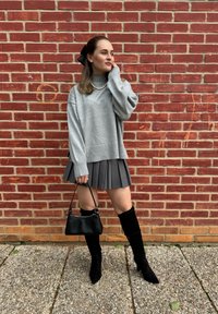 Γκρι oversized πουλόβερ, πλισέ γκρι φούστα, μαύρες ψηλές μπότες και μαύρη τσάντα κατά μήκος ενός τοίχου από τούβλα. Υφές και προσεγμένος σχεδιασμός.