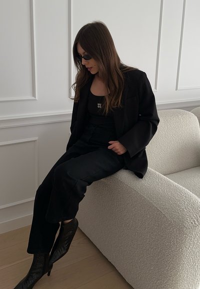 Blazer negro sobre una camiseta sin mangas negra con el logo "HUGO", combinado con pantalones cortos negros y botas de tacón negras texturizadas, sentado en un sofá claro.