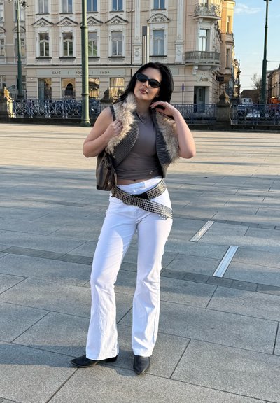 Mujer con gafas de sol, chaleco gris sin mangas con ribete de piel, top gris, pantalones blancos y botas negras posando en una plaza de la ciudad con edificios históricos detrás.