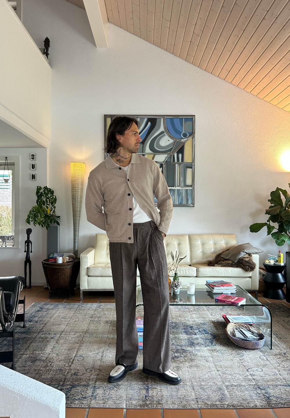 Lys beige cardigan med mørke knapper, kombinert med brune rutete bukser. Glatt stoff, avslappet passform, står i et moderne stue.
