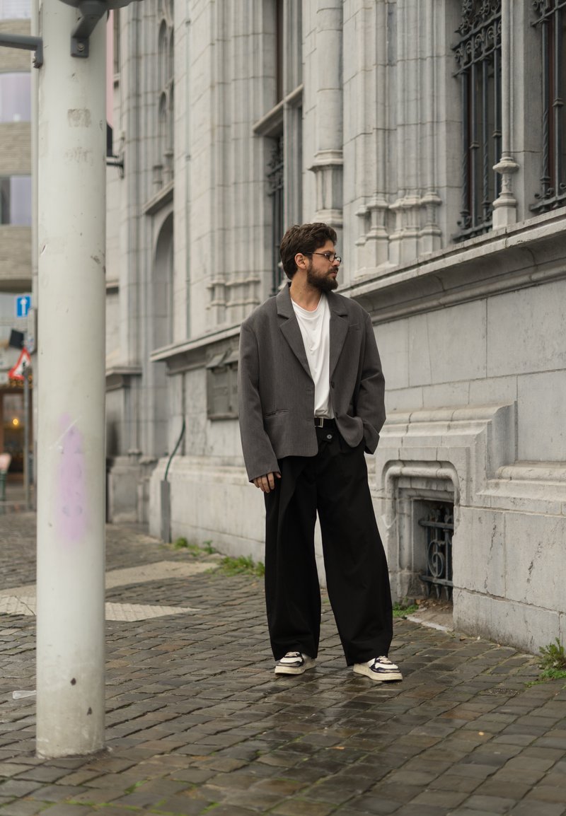Blazer grigio oversize sopra una t-shirt bianca, abbinato a pantaloni neri a gamba larga e sneakers beige, in piedi su una strada di ciottoli.
