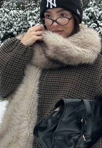 Manteau pied-de-poule marron avec un col en fausse fourrure beige, sac à main noir et bonnet noir, portant des lunettes rondes et une main ajustant le col.