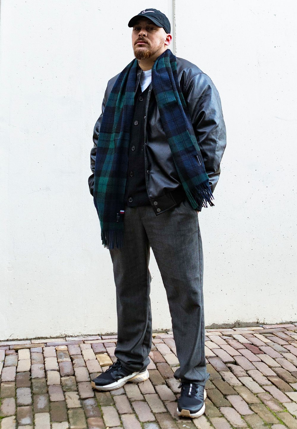 Fekete bőrkabát, tengerészkék kockás sál, szürke mellény, szürke nadrág és fekete sportcipő. Egy textúrált fehér falnak támaszkodik, macskaköves úton.