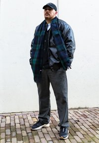 Fekete bőrkabát, tengerészkék kockás sál, szürke mellény, szürke nadrág és fekete sportcipő. Egy textúrált fehér falnak támaszkodik, macskaköves úton.