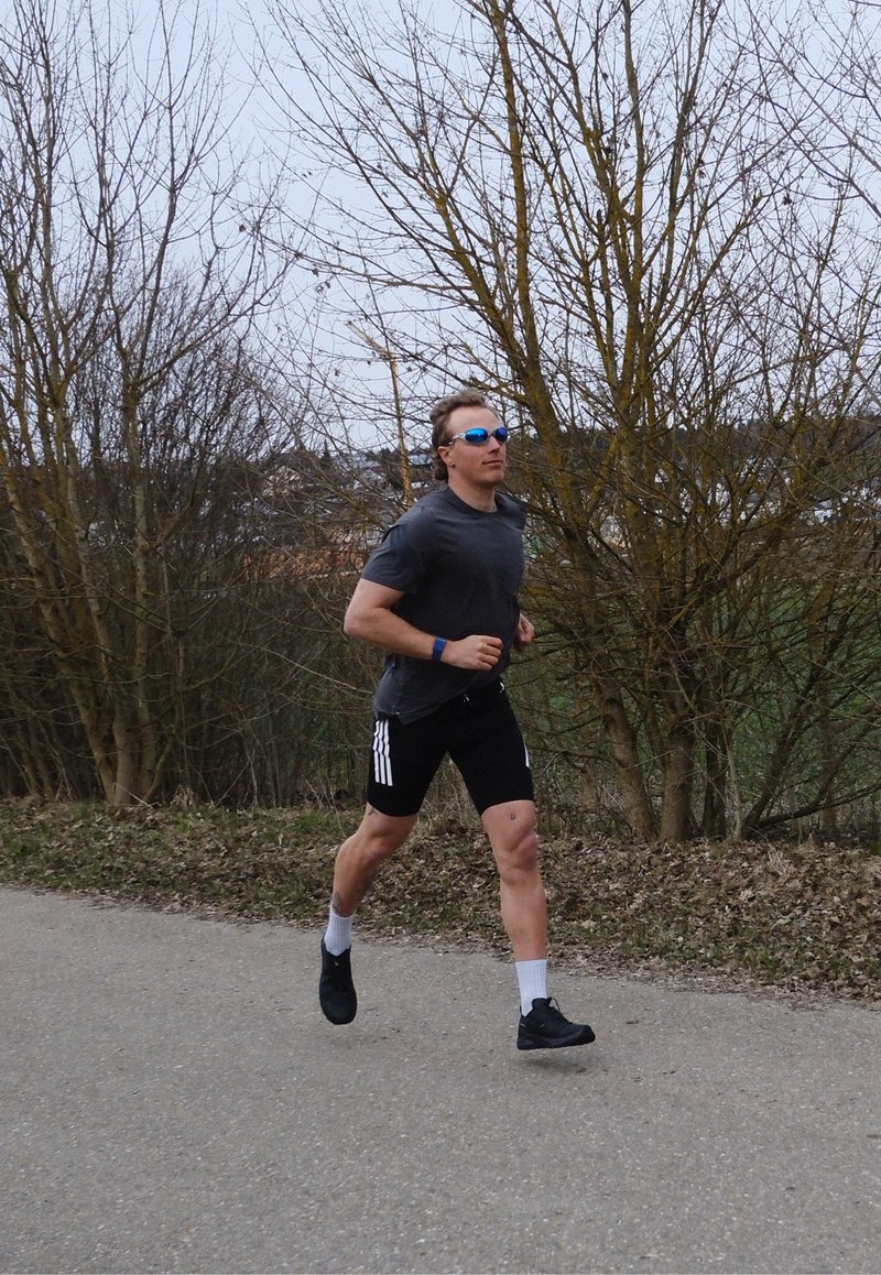 Man wearing black shorts, gray shirt, and blue sunglasses jogging on a paved path near leafless trees in early spring.