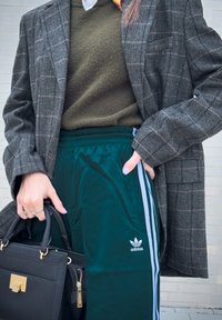 Grå hundetann-blazer, olivengrønn genser, grønne treningsbukser med hvite sidestriper, svart håndveske med gullbeslag, holdt i hånden.