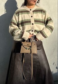 Hrubý krémový cardigan so zelenými a hnedými pruhmi, zapínanie na gombíky, v kombinácii s tmavou koženou sukňou a malou béžovou kabelkou s kovovými detailmi.