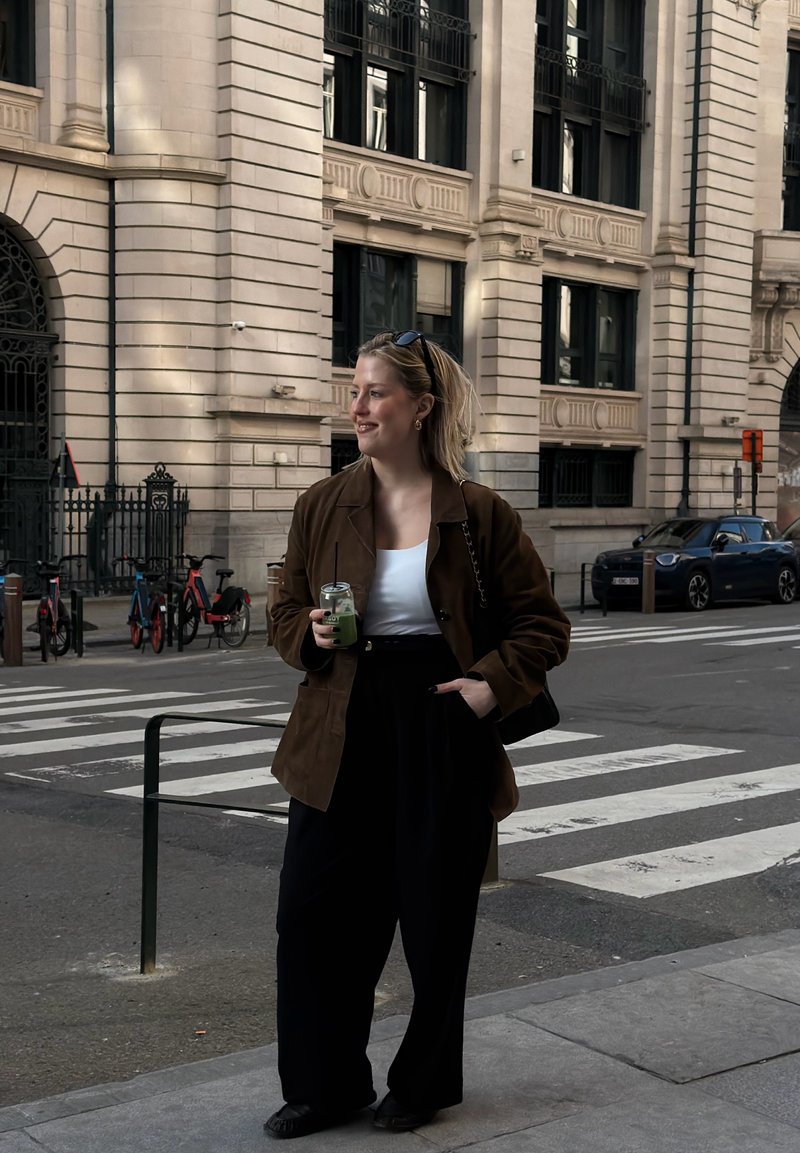 Jeune femme tenant un smoothie vert, debout près d'un passage piéton en ville, portant un blazer marron, un haut blanc, un pantalon noir, avec des lunettes de soleil sur la tête.