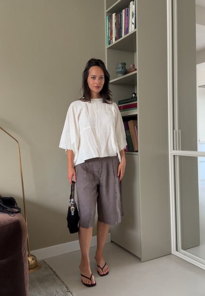 Top de lino blanco y holgado con cuello redondo, combinado con shorts de lino grises. Bolso negro con cadena y sandalias tipo flip-flop.