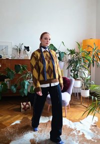 Γυναίκα στέκεται μέσα σε εσωτερικό χώρο φορώντας καφέ και κίτρινο πουλόβερ με σχέδιο ρόμβου, λευκό πουκάμισο, σκούρο μπλε παντελόνι, μπλε παπούτσια, μπλε κασκόλ, περικυκλωμένη από φυτά και διακοσμητικά αντικείμενα.