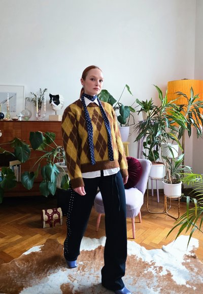Mujer está de pie en interiores con un suéter marrón y amarillo de rombos, camisa blanca, pantalones azul marino, zapatos azules y bufanda azul, rodeada de plantas y decoración.