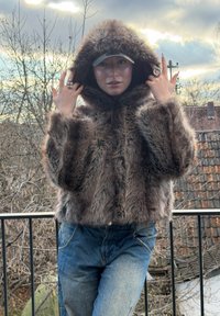Faux fur cropped jacket in shades of brown, featuring a hood and wide sleeves, paired with blue denim trousers and a cap.