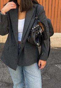 Szürke kockás blézer laza szabással, fehér trikóval és világoskék farmerrel párosítva. Texturált fekete kézitáskát tart ezüst díszítésekkel.