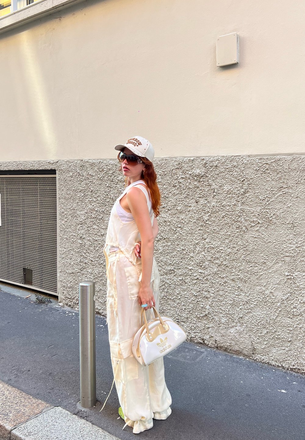Μακρύ φόρεμα σε σατέν κρέμα απόχρωσης με ρυθμιζόμενες  straps, συνδυασμένο με μια λευκή τσάντα χειρός με χρυσές λεπτομέρειες. Το μοντέλο φοράει ένα μπεζ καπέλο και γυαλιά ηλίου.