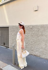 Μακρύ φόρεμα σε σατέν κρέμα απόχρωσης με ρυθμιζόμενες  straps, συνδυασμένο με μια λευκή τσάντα χειρός με χρυσές λεπτομέρειες. Το μοντέλο φοράει ένα μπεζ καπέλο και γυαλιά ηλίου.