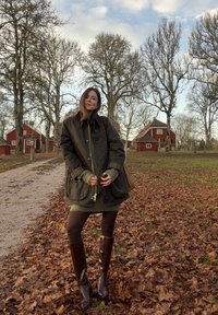 Tmavě zelená nepromokavá bunda se dvěma předními kapsami, v kombinaci s pletenými zelenými šaty, hnědými punčocháči a vysokými hnědými koženými botami.