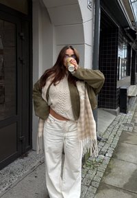 Ylisuuri oliivinvihreä takki kerroksena kermaisen neulotun villapaidan päällä, valkoiset leveälahkeiset housut ja beige ruudullinen huivi. Kädessä take away -kahvikuppi.