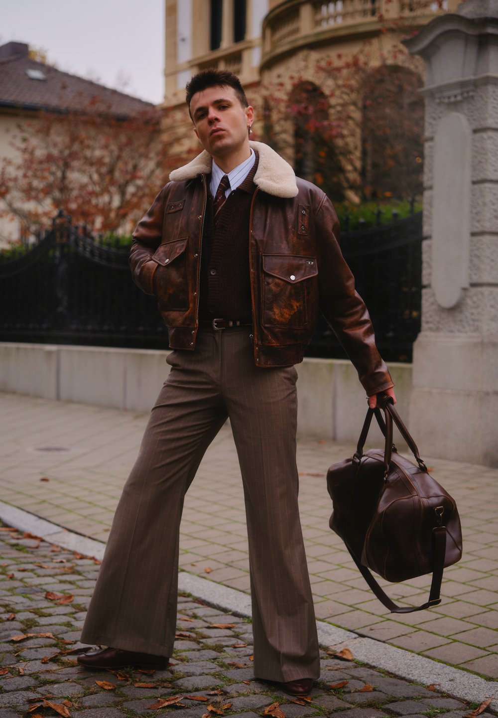 Brun skinnjakke med en krage i pelskledd skinn, rutete brune bukser og en brun skinnreisetaske. Stylet med hvit skjorte og slips.