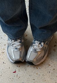 Zapatillas de malla plateadas y grises con un cuello acolchado, cordones y una suela de goma blanca, usadas con vaqueros de denim azules.