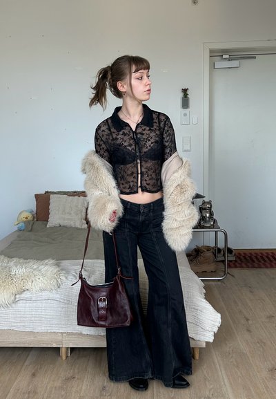 Joven con coleta que lleva un top de encaje negro, mangas de pelo, jeans acampanados, sosteniendo un bolso marrón, de pie en una habitación con cama y zapatos cerca.