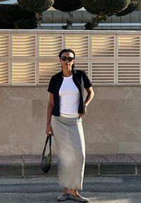 Μαύρο κοντομάνικο μπουφάν πάνω από λευκό πουκάμισο, γκρίζα μάξι φούστα, μαύρη τσάντα και μαύρες μονοτζιανικές μπαλαρίνες σε καμβά με υφή και πράσινα φυτά.