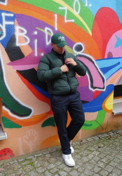 Chaqueta de plumas verde con un alto cuello y cremallera frontal, pantalones azul marino y zapatillas blancas, frente a un fondo de grafitis colorido.