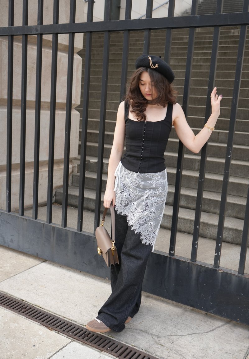 Młoda kobieta w czarnej bluzce, spódnicy z koronkową nakładką, szerokich spodniach i berecie, trzymająca torebkę, stojąca przy czarnym żelaznym ogrodzeniu i kamiennych schodach.