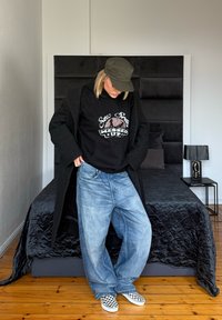 Manteau noir oversize, sweat à capuche noir avec motif graphique, jean large bleu et baskets slip-on à damier. Tenue simple et décontractée dans une chambre.