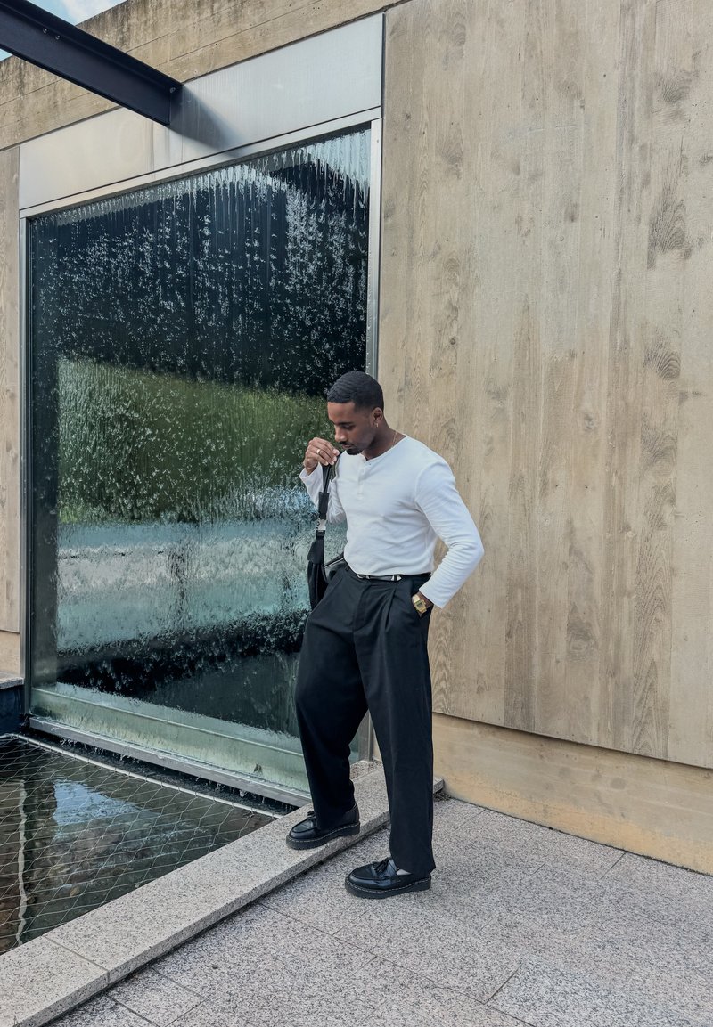 Pantaloni larghi neri e una maglietta a maniche lunghe bianca stile henley, in piedi accanto a una fontana con un muro testurizzato sullo sfondo.