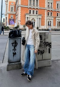 Trench-coat beige, pull blanc, jean large bleu clair délavé, baskets noires avec lacets blancs, et sac à main noir, à côté d'une boîte utilitaire marquée de graffitis.