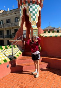 Colourful tiled steps leading to a textured red architectural structure. A female figure wearing a red sweatshirt, black shorts, and grey shoes.