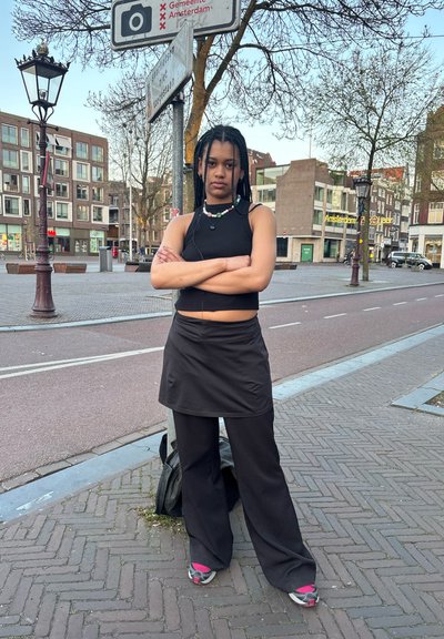 Top sin mangas negro, falda negra y pantalones anchos negros. La joyería incluye un collar multicolor. Entorno urbano con árboles y edificios.
