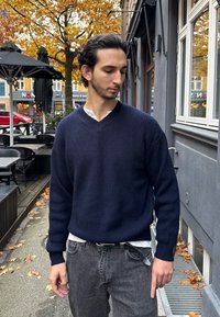 Tumedast meresinise ribitatud kudumisega sviiter V-kaelusega, kantud valge kraega särk üle ning hallide teksadega. Väljas linnakeskkonnas sügislehtede seas.