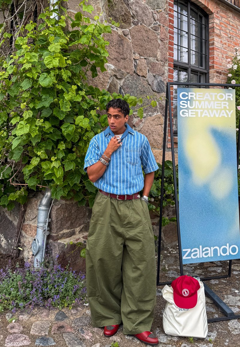 Personne portant un pantalon vert large, une chemise à rayures bleues et des chaussures rouges, debout à côté d'une plante, près d'un panneau de l’événement Zalando et d’un sac avec une casquette rouge.