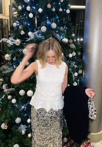 Femme portant un haut blanc sans manches et une jupe à sequins, debout devant un sapin de Noël décoré, tenant un manteau et un petit sac à main.