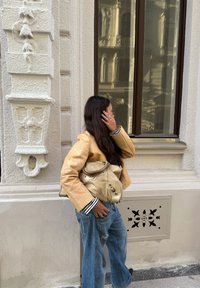 Gelber, strukturierter Blazer über einem gestreiften Hemd, dazu ausgewaschene blaue Jeans. Eine beige Ledertasche mit Akzenten. Hintergrund aus einer strukturierten Wand und einem Fenster.