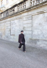 Persoon die over een grindpad naast een grote stenen muur loopt, gekleed in een donker oversized blazer en broek, en een zwarte geweven handtas draagt.