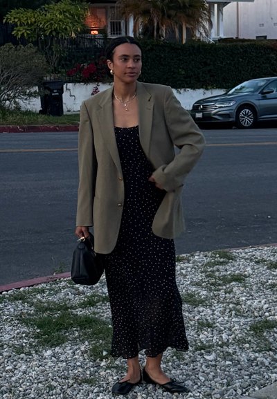 Blazer verde oliva de gran tamaño sobre un vestido negro con lunares, complementado con un pequeño bolso negro y zapatos planos negros. Fondo de grava.