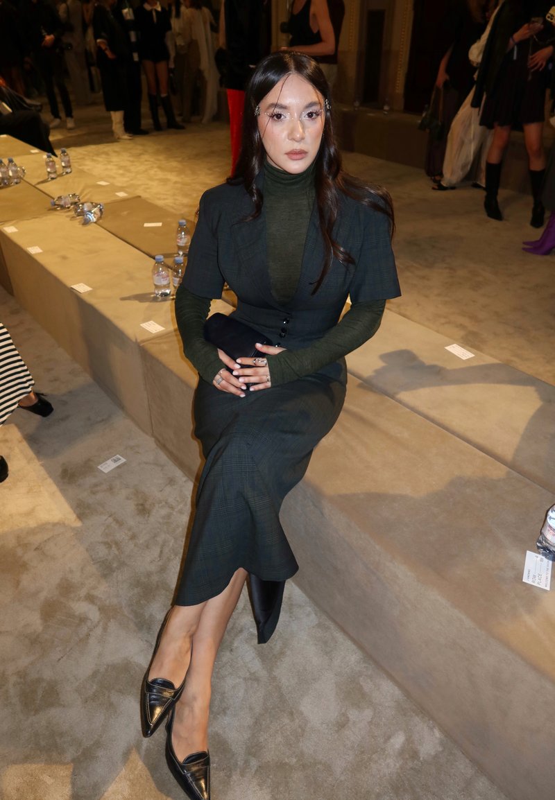 Femme en robe sombre à couches et mocassins noirs assise sur un banc beige lors d'un événement en intérieur avec des personnes et des bouteilles d'eau en arrière-plan.