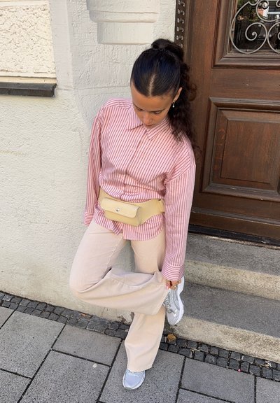Camisa de rayas rosa y blanca con cuello, bolsa de cinturón beige, pantalones anchos de color crema y zapatillas gris claro, de pie en unos escalones de piedra.