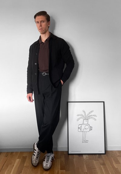 Cárdigan negro sobre un polo marrón, combinado con pantalones negros. Zapatillas gris con rayas blancas. Obra de arte enmarcada en el suelo al lado.