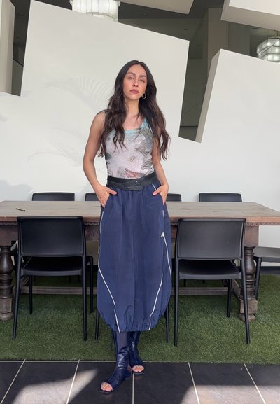 Mujer con cabello largo y oscuro, de pie con las manos en los bolsillos, vistiendo un top sin mangas con estampado, falda azul y botas abiertas en una habitación moderna.