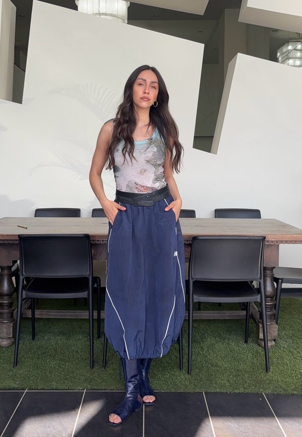 Frau mit langen dunklen Haaren steht mit den Händen in den Taschen, trägt ein ärmelloses bedrucktes Top, einen blauen Rock und offene Stiefel in einem modernen Raum.