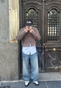 Camisola curta castanha de malha tricotada, camisa branca abotoada, jeans largos azul claro, boné preto com letras brancas, sapatos pretos com detalhes brancos.