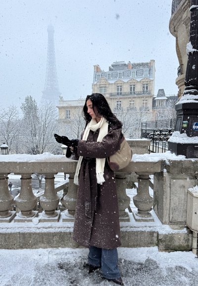 Mujer con abrigo marrón, bufanda blanca y guantes negros de pie en una calle nevada de París con la Torre Eiffel y un edificio histórico al fondo.