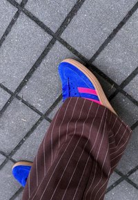 Sneakers in suede blu con strisce rosa, pantaloni a righe marroni e suola in gomma, su una superficie di piastrelle grigie.