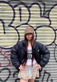 Casaco puffer preto, top de alças branco com grafismo, saia mini xadrez e um boné camuflado. Cabelo comprido com mechas vermelhas em um fundo de graffiti.