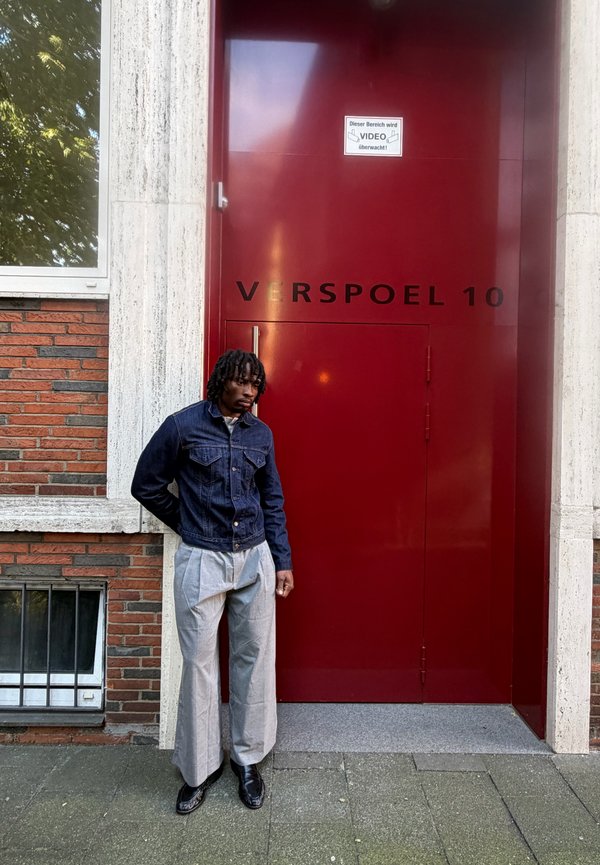 Hombre con chaqueta vaquera y pantalones grises está de pie con las manos detrás de la espalda frente a una gran puerta roja con la marca "VERSPOEL 10".