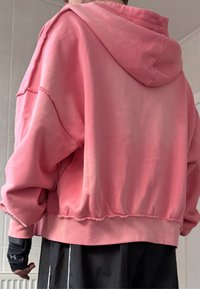 Roze cropped hoodie van zachte stof, met een capuchon, rafelige randen en een losse pasvorm. G gedragen over zwarte sportshorts met witte strepen.